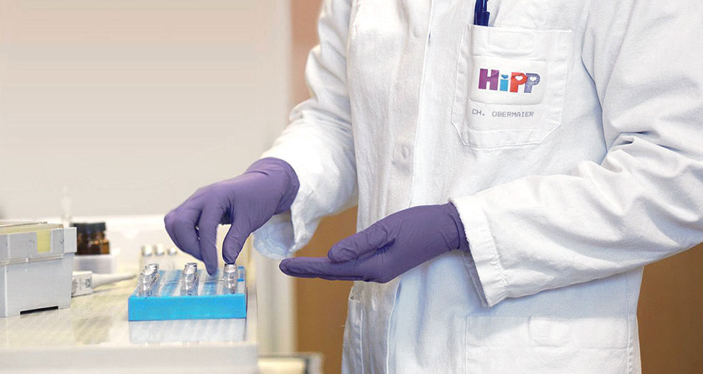 A person in a white lab coat with the logo "HiPP" and the name "CH. OBERMAIER" is wearing purple gloves and handling a blue rack with multiple small containers.